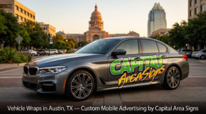Professional product photography of a gray BMW 5 Series sedan featuring custom vehicle wraps with the Capital Area Signs logo. The wrap displays "CAPITAL" in bold green block letters and "Area Signs" in cursive orange-yellow letters on the side of the car. The vehicle is parked at an angle on a brick-patterned sidewalk. Late afternoon golden hour lighting with warm natural light creating subtle reflections. Urban Austin setting with the iconic Texas State Capitol building and Frost Bank Tower clearly visible in the background, along with brick pavement and city buildings. Sharp focus on the wrapped BMW with shallow depth of field bokeh effect. Dark banner at bottom with white text overlays reading "Vehicle Wraps in Austin, TX — Custom Mobile Advertising by Capital Area Signs." High-quality commercial automotive photography showcasing premium vehicle wrap services in Austin, Texas with recognizable local landmarks.